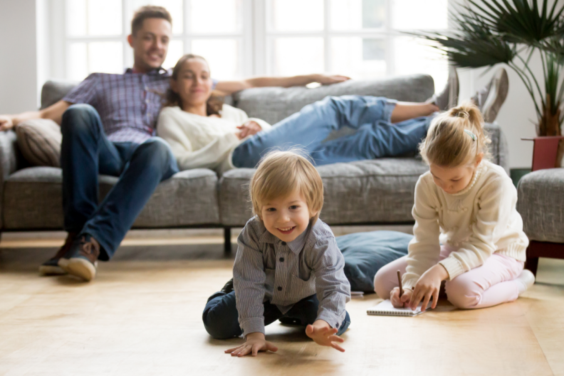 Ein junges Paar sitzt vor einem Fenster auf seinem Sofa, davor spielen seine beiden Kinder auf dem Fußboden.