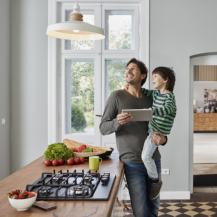 Ein junger Vater steht mit seinem Sohn auf dem Arm in der Küche und kontrolliert den Energieverbrauch über ein Tablet.