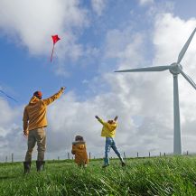 Ein Vater lässt mit seinen zwei Kindern auf einer Wiese Drachen steigen. Im Hintergrund stehen Windenergieanlagen.