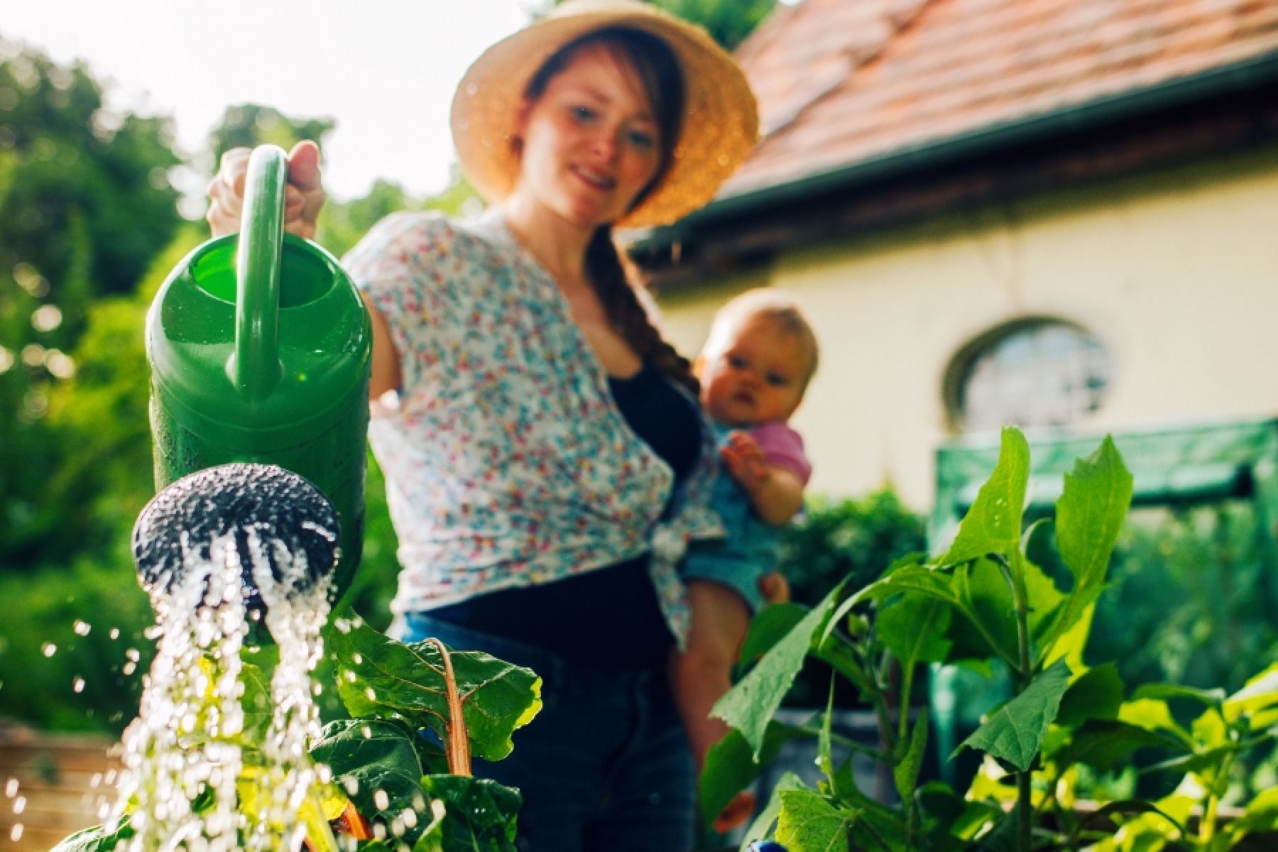 Eine Frau hat ihr Baby auf dem Arm und gießt ein Beet.