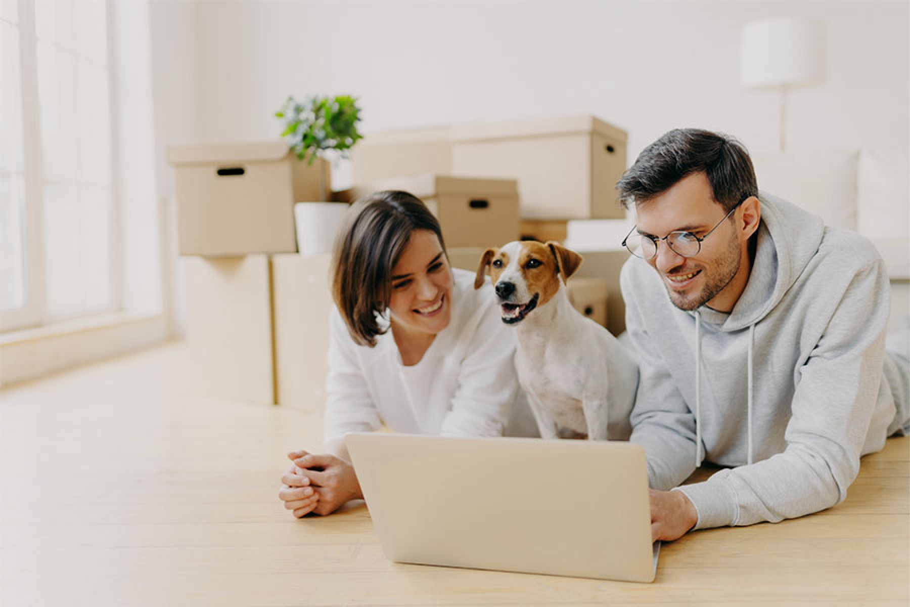 Ein junges Paar liegt mit seinem Hund auf dem Wohnzimmerfußboden vor gepackten Umzugskartons.