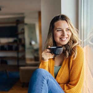 Eine junge Frau sitzt auf der Fensterbank und trinkt eine Tasse Kaffee. 