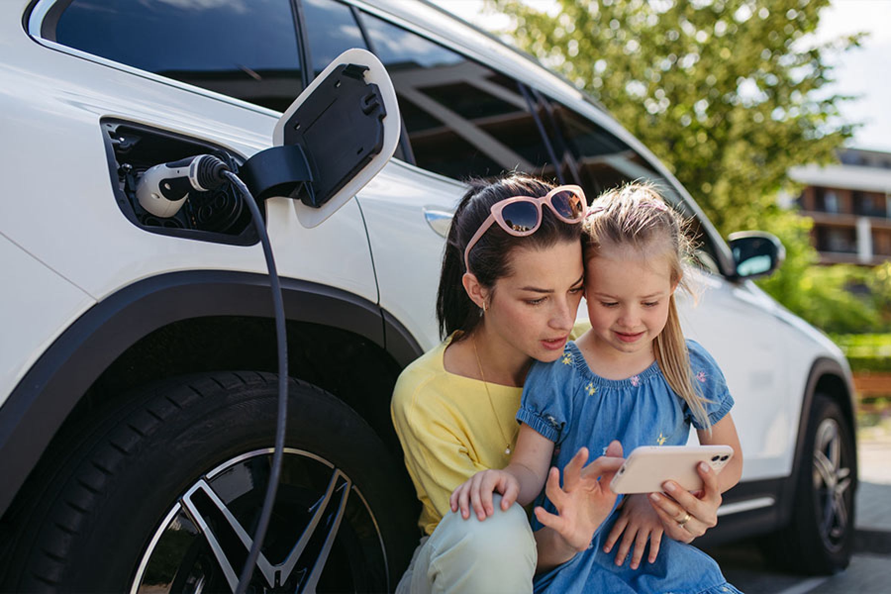 Eine Mutter und ihre kleine Tochter nutzen ein Smartphone während das E-Auto aufgeladen wird.  