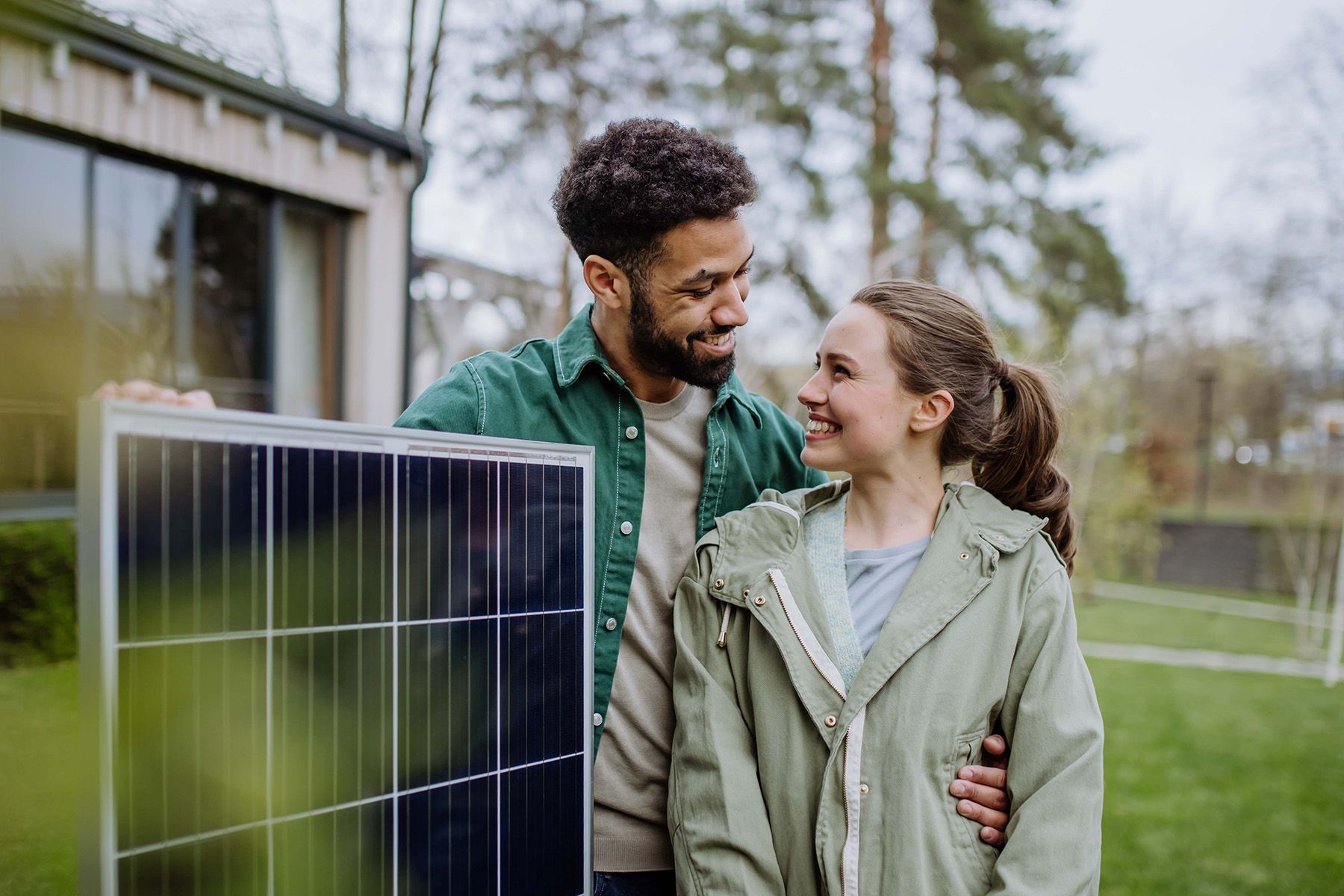 Ein junges Paar mit einem Solarmodul vor einem Haus.