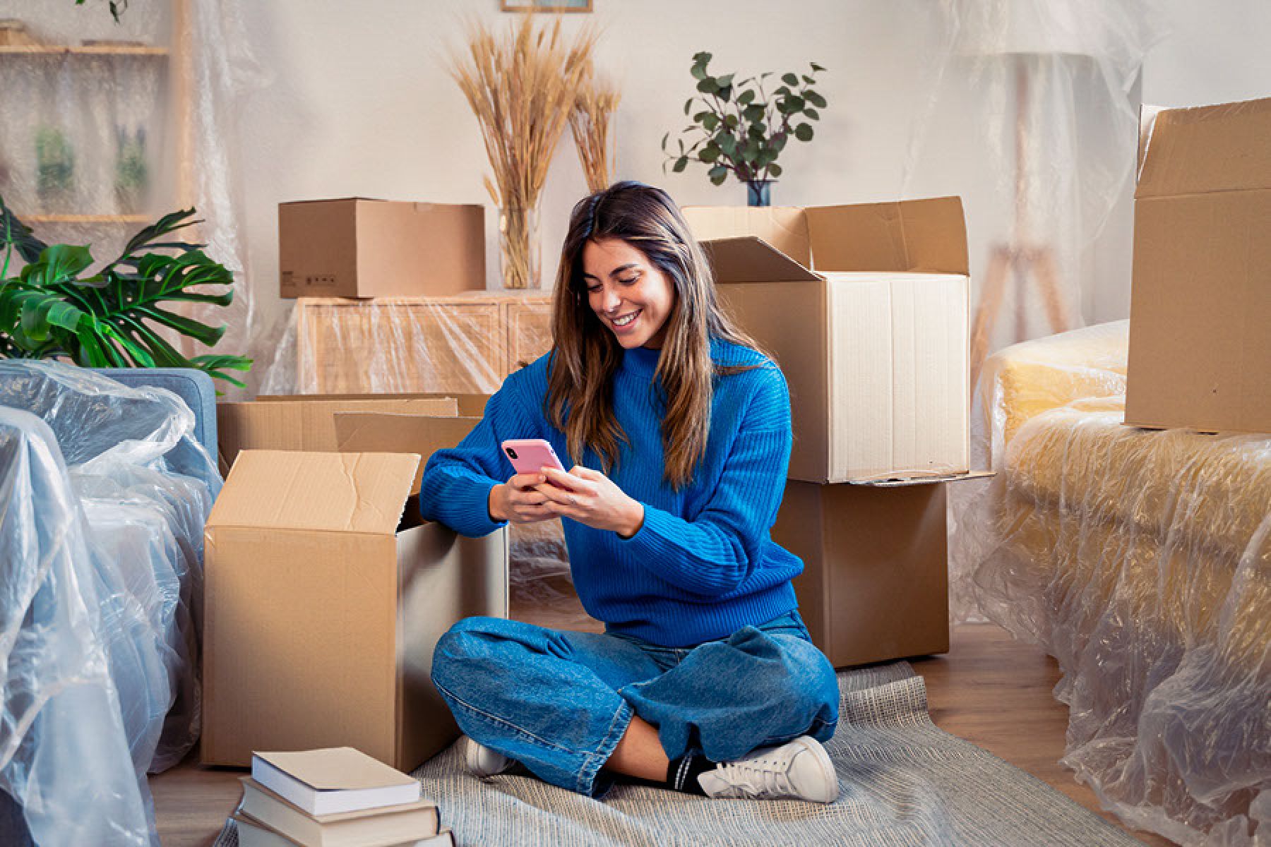 Eine junge Frau sitzt im Wohnzimmer auf dem Fußboden inmitten von Um-zugskartons.