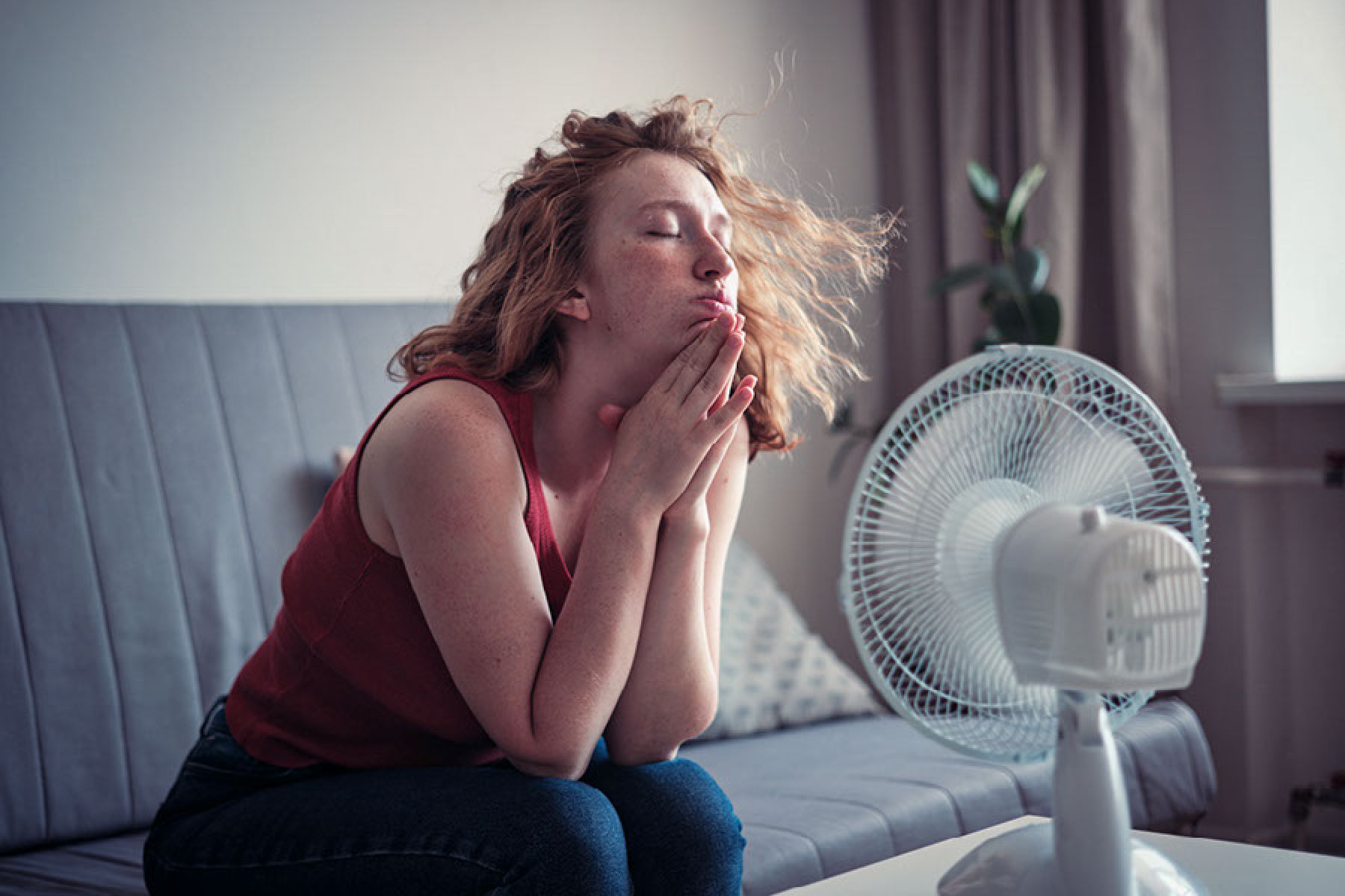 Eine junge Frau sitzt erschöpft auf dem Sofa und lässt sich von einem Tischventilator abkühlen.  
