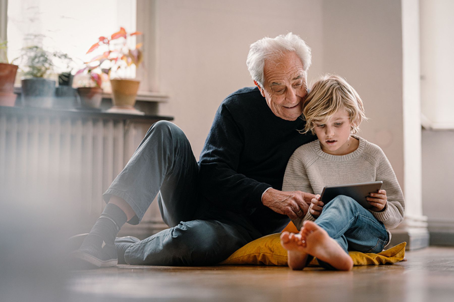 Ein Großvater sitzt mit seinem Enkel auf einem Sitzkissen auf dem Boden, im Hintergrund ist ein Heizkörper zu sehen.