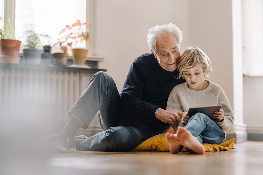 Wohlige Wärme dank nachhaltiger Fernwärme: Großvater und Enkel nutzen zusammen ein Tablet und sitzen dabei auf dem Fußboden.