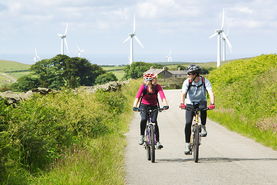 Zwei Radfahrer fahren mit E-Bikes durch die Natur. Im Hintergrund sind Windenergieanlagen zu sehen. 