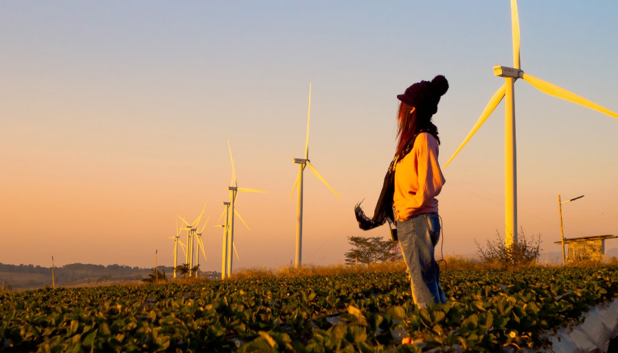 Eine junge Frau steht in einem Feld mit Windenergieanlagen und schaut in die Ferne.
