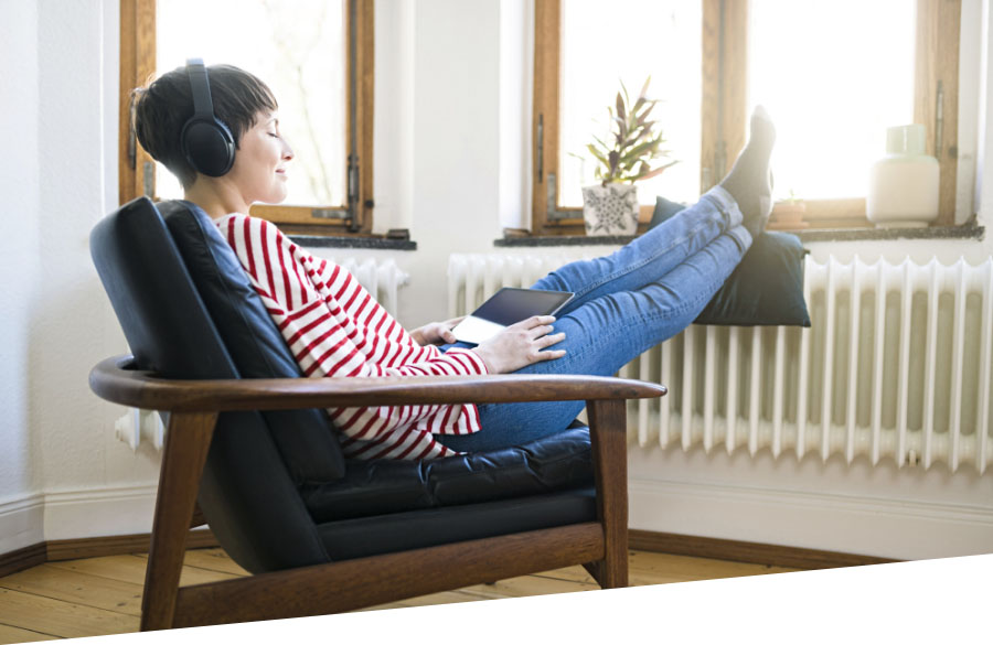 Eine optimal eingestellte Heizung und dichte Fenster helfen beim Energie sparen. Eine junge Frau sitzt in einem Sessel, hört Musik und hat ihre Füße auf der Heizung abgelegt.