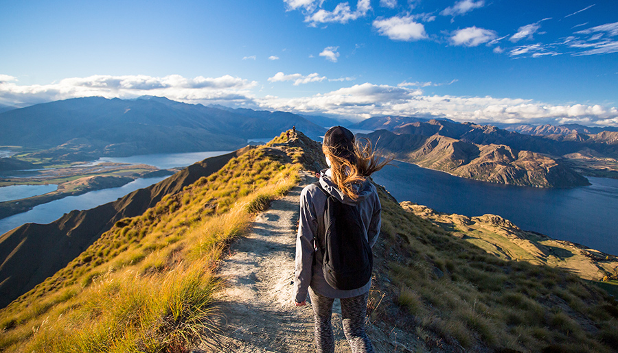 Eine junge Frau wandert auf einem Gipfelpfad, vor ihr liegt ein Panorama von Bergen und Fjorden.