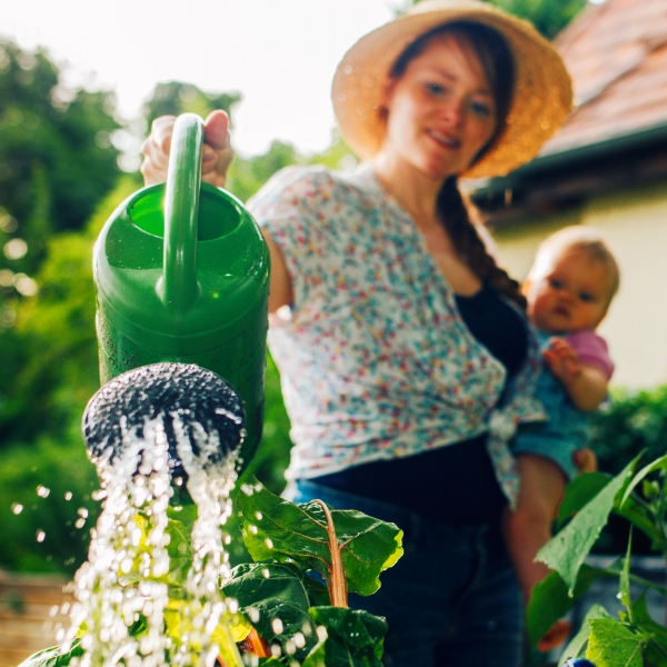 Eine Frau hat ihr Baby auf dem Arm und gießt ein Beet.