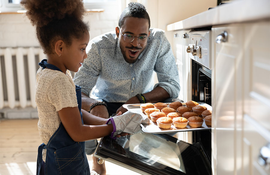 Ein Mädchen zieht ein Blech mit gebackenen mit Muffins aus dem Ofen. Ihr Vater schaut zu.
