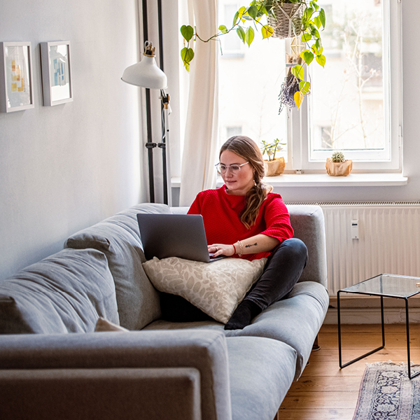 Eine junge Frau sitzt auf ihrem Sofa und nutzt ihren Laptop.