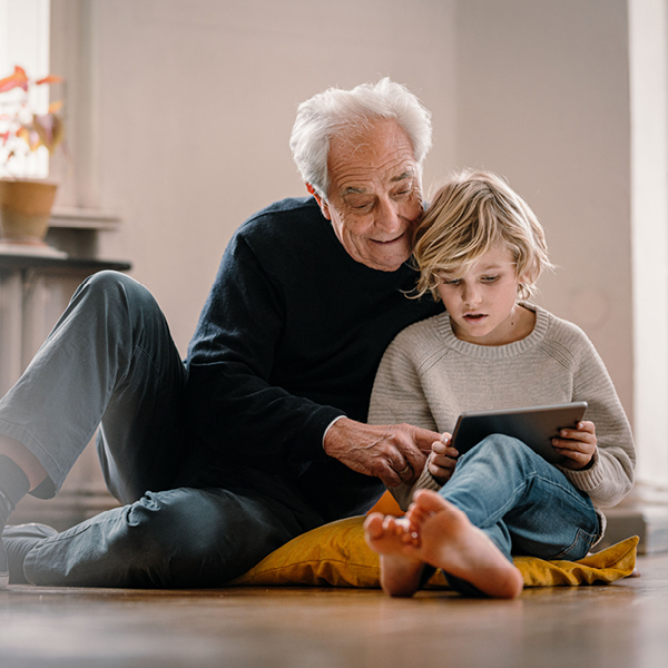Ein Großvater sitzt mit seinem Enkel auf einem Sitzkissen auf dem Boden, im Hintergrund ist ein Heizkörper zu sehen.
